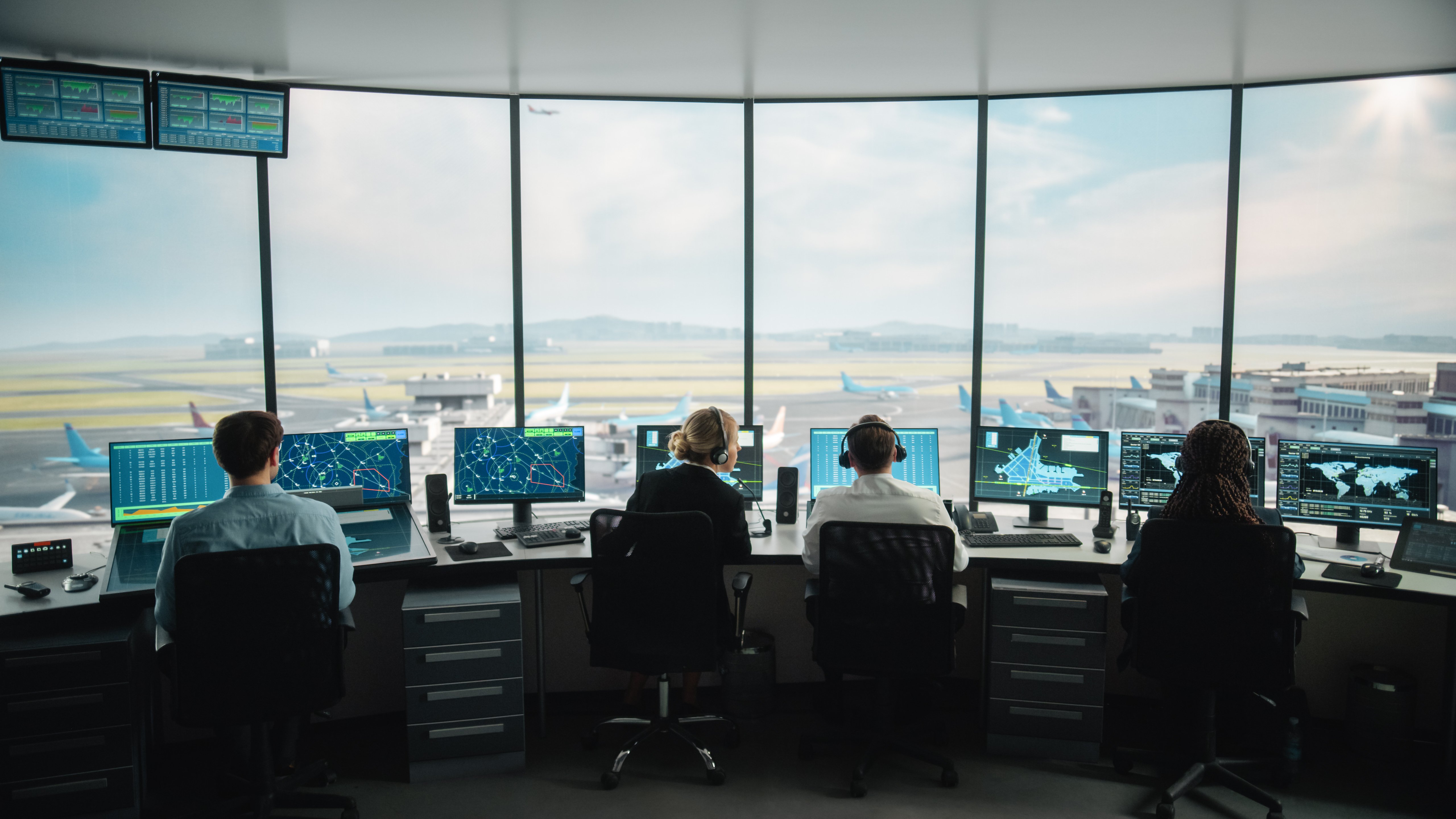 Quatro controladores de tráfego aéreo estão sentados em estações de trabalho em uma torre de controle de aeroporto, cada um monitorando várias telas que exibem radares, rotas de voo, mapas de aeroportos e dados globais. Grandes janelas panorâmicas oferecem uma visão clara da pista do aeroporto, dos aviões em taxiamento e dos edifícios do terminal. A equipe coordena os movimentos das aeronaves em um ambiente seguro e de alta tecnologia