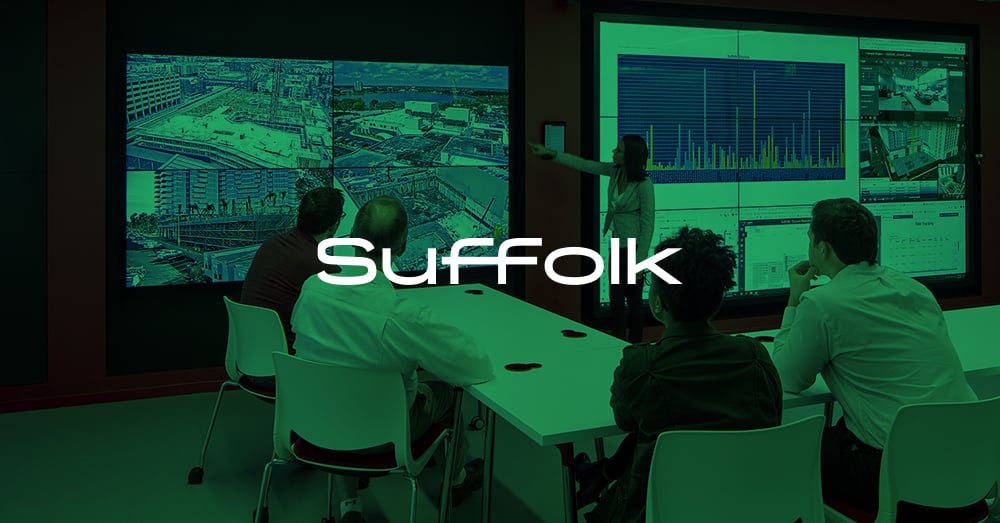 Empregado em pé apresenta-se usando parede de vídeo, 4 empregados vêem apresentação sentados em uma mesa em Suffolk's L.A. Smart Lab com sobreposição verde e logotipo