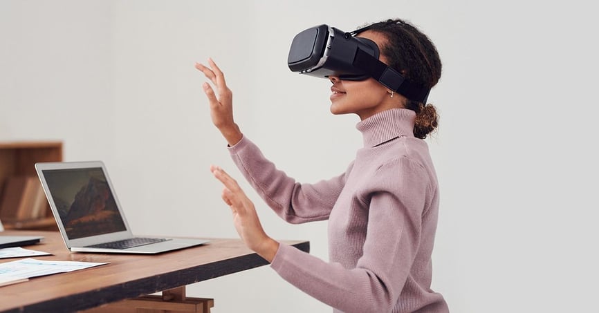 Mulher usando um fone de ouvido VR sentada em sua mesa que tem notas e um laptop