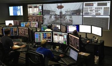 Uma sala de controle com trabalhadores em suas estações de trabalho e uma parede de vídeo exibindo filmagens e dados ao vivo