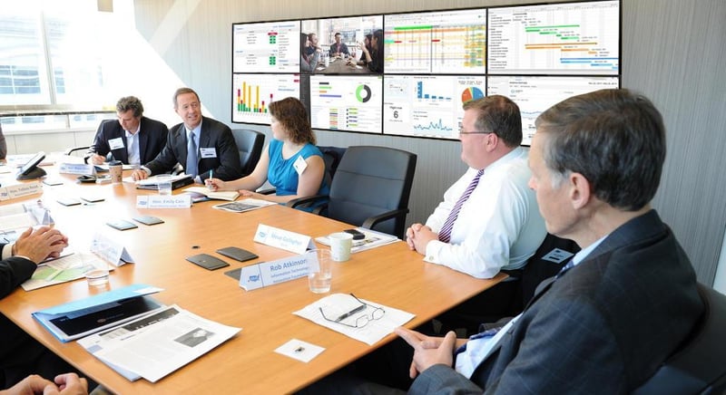 5 funcionários têm uma discussão enquanto estão sentados à mesa em uma sala de reunião com uma parede de vídeo exibindo visualizações de dados