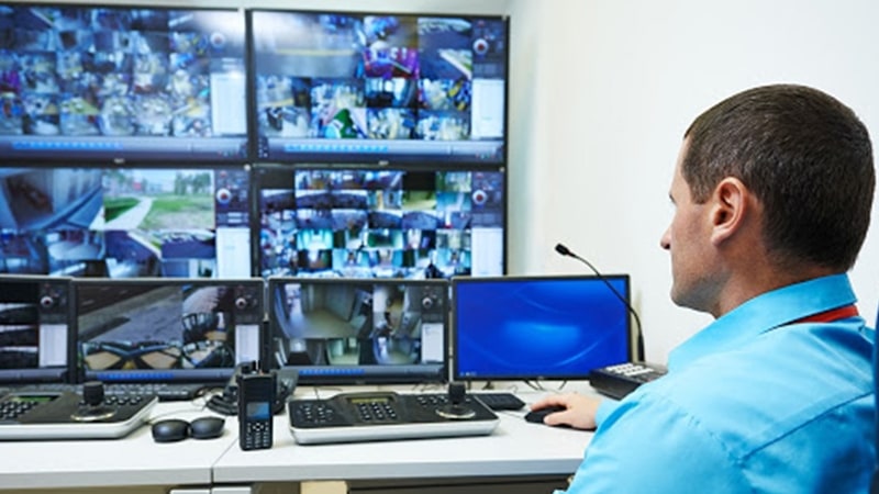Homem no centro de controle de segurança do campus monitorando o campus através de uma parede de vídeo exibindo filmagens ao vivo da câmera