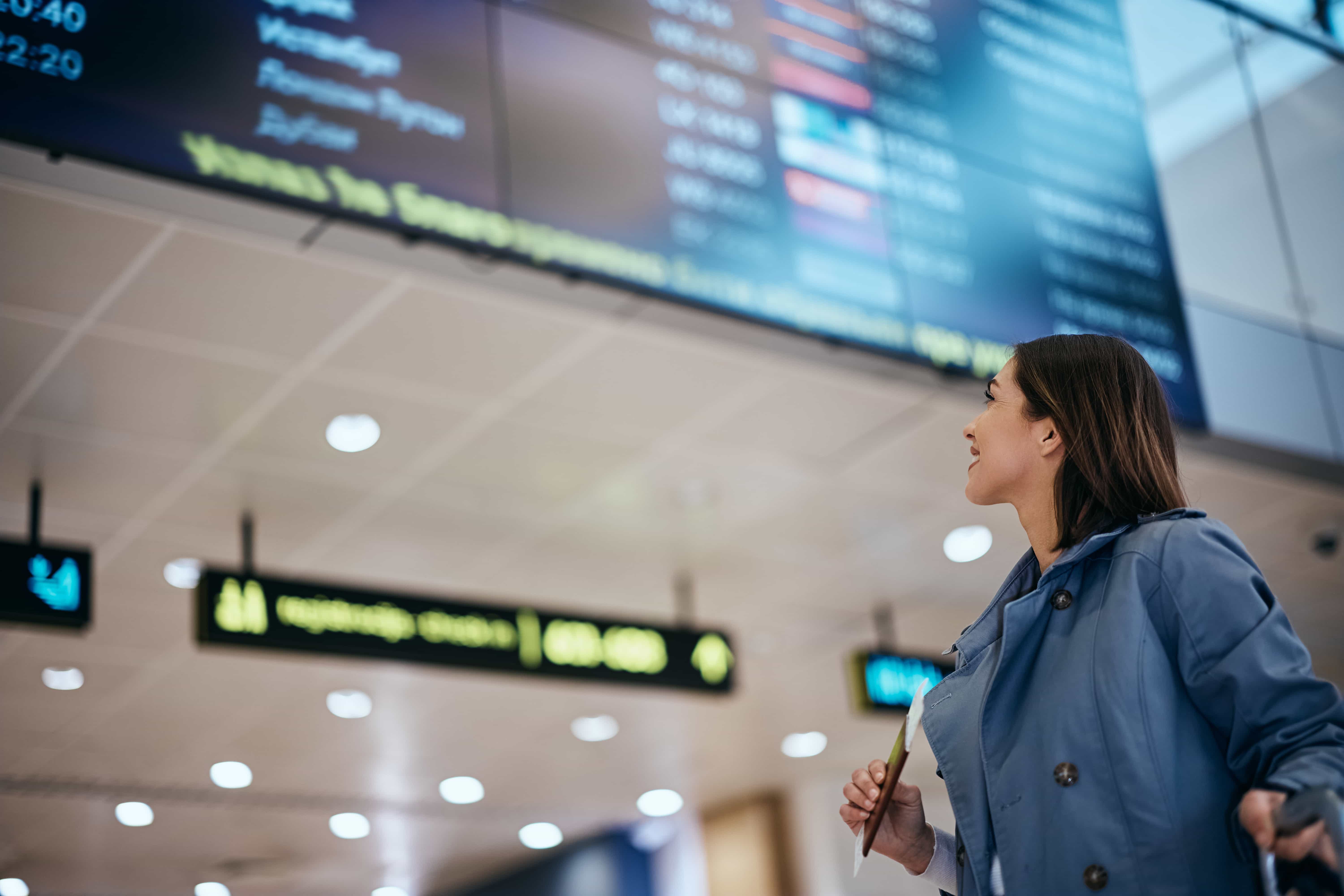  Uma mulher de jaqueta azul clara e camisa branca olha para um grande quadro de partidas em um aeroporto. Ela está sorrindo levemente e segurando o que parece ser um passaporte ou passagem em sua mão direita. O quadro de partidas, que está fora de foco, exibe várias informações de voo. O teto do aeroporto com luzes também é visível ao fundo.