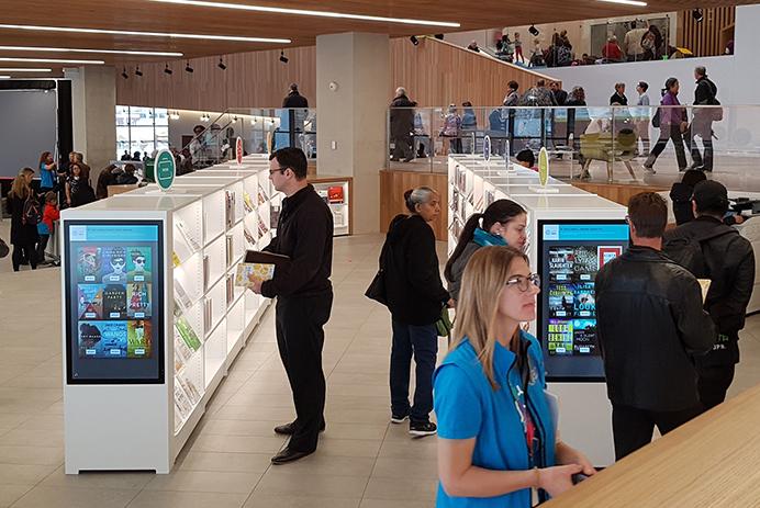  Muitas pessoas em uma biblioteca andam e vêem prateleiras com sinalização digital.