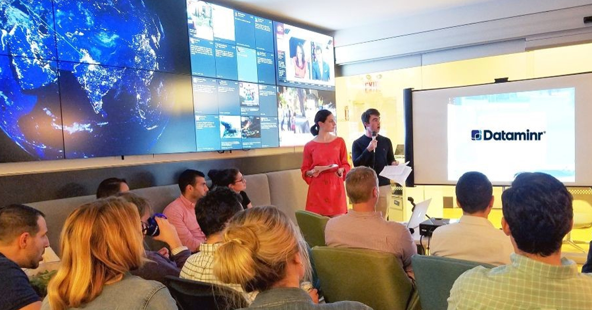 Funcionários da Dataminr em uma sala de reuniões sentados em frente a um projetor, e ao lado de uma parede de vídeo com mídias sociais e notícias atualizadas