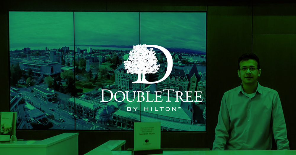 DoubleTree by Hilton recepcionista e 3 painéis de vídeo atrás dele exibindo pontos de referência de Victoria, Canadá, com sobreposição verde e logotipo