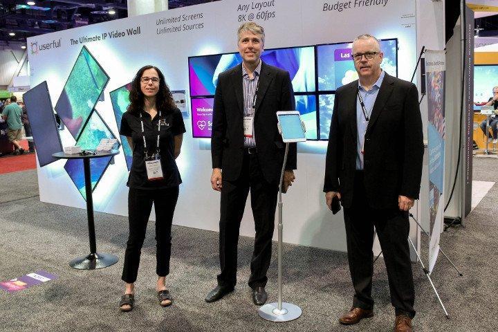 3 Empregados usuários no Infocomm 2018, em frente ao estande do usuário AV-over-IP com uma parede de vídeo