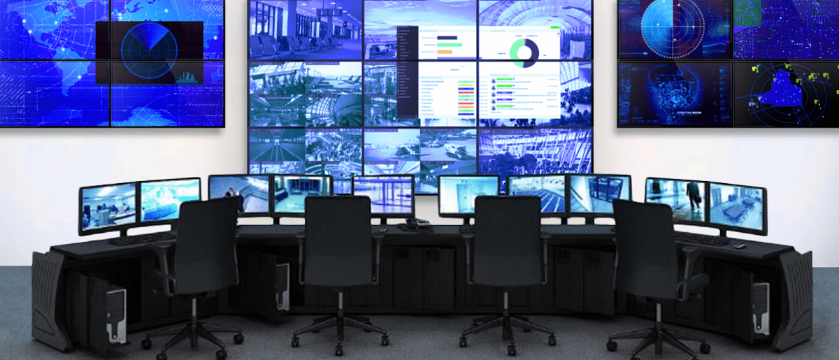 Sala de controle vazia com 4 estações de trabalho e 3 paredes de vídeo exibindo imagens de câmeras de segurança ao vivo e painéis de dados