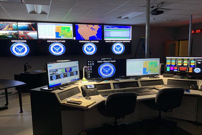 Sala de controle EMA vazia, com estação de trabalho, e uma parede de vídeo exibindo fotografia, dados e mapas