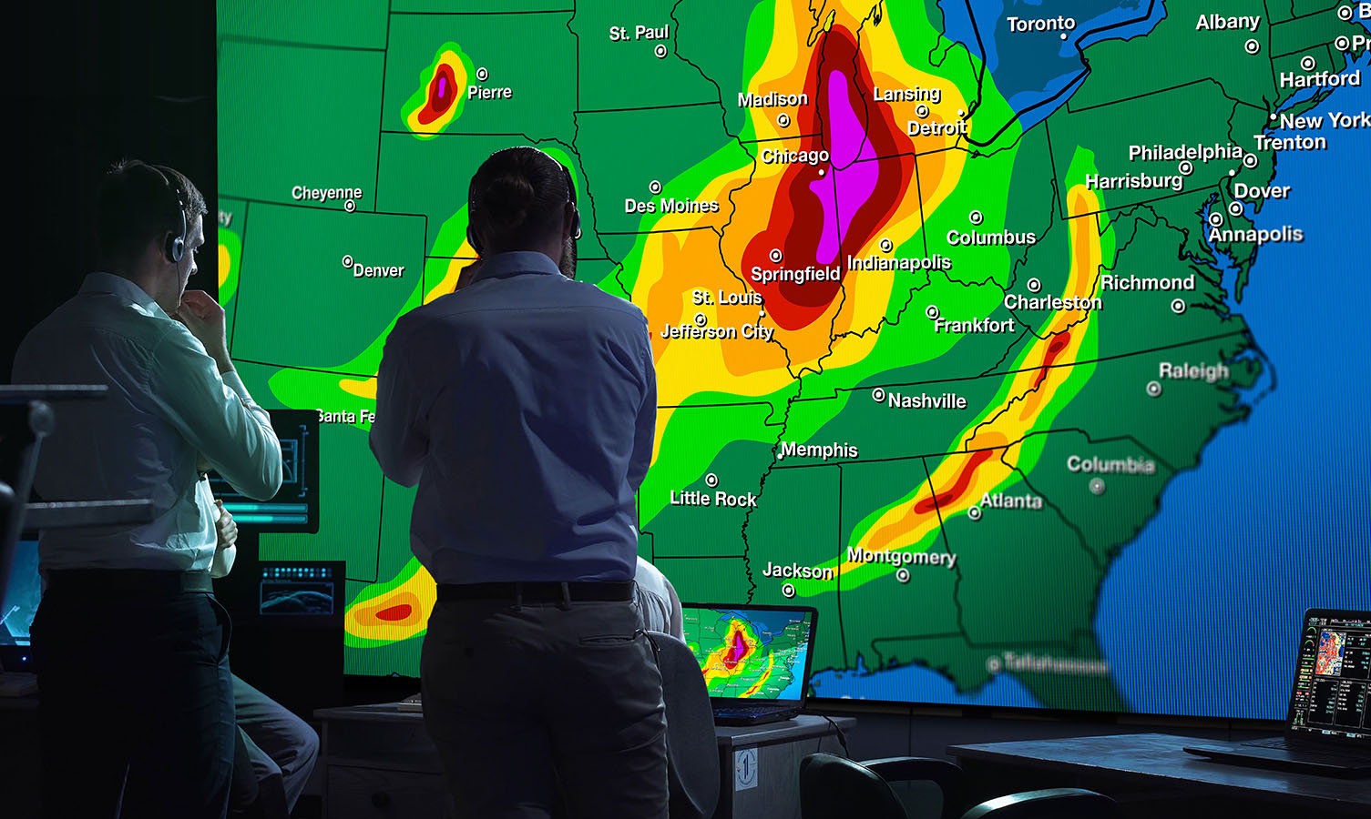 2 homens em uma sala de controle de monitoramento meteorológico, olhando para uma grande parede de vídeo exibindo um mapa meteorológico dos EUA