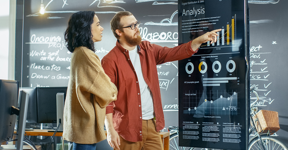 Um homem e uma mulher discutem um painel de dados em uma tela de TV, com um quadro-negro com detalhes do projeto por trás deles