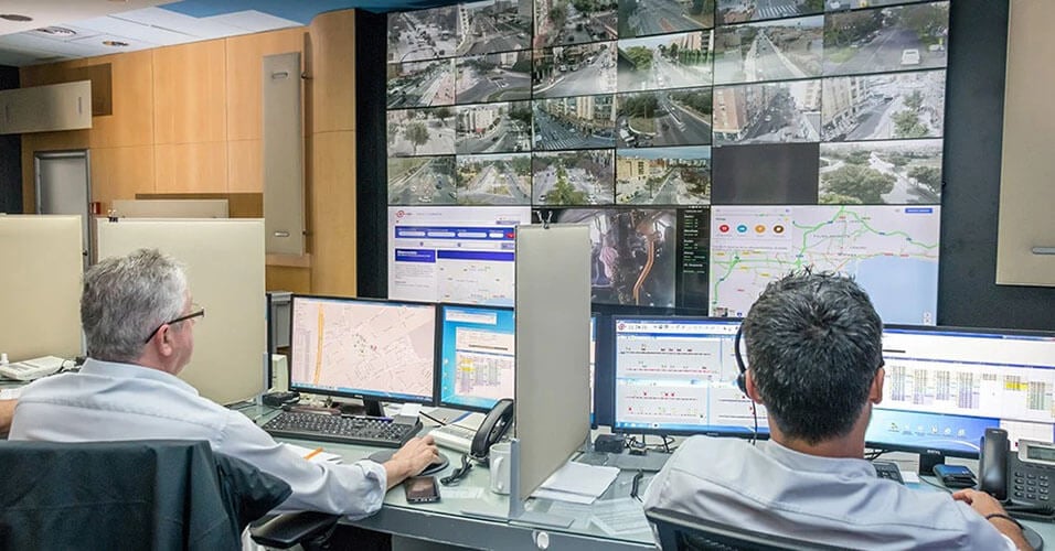 Dois funcionários da EMT monitorando operações de trânsito através de suas estações de trabalho e uma parede de vídeo exibindo imagens de câmeras ao vivo, mapas de trânsito e sites