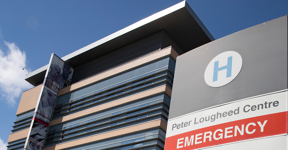 Uma foto da placa de emergência do Peter Lougheed Center Hospital, e do edifício do hospital contra um céu azul